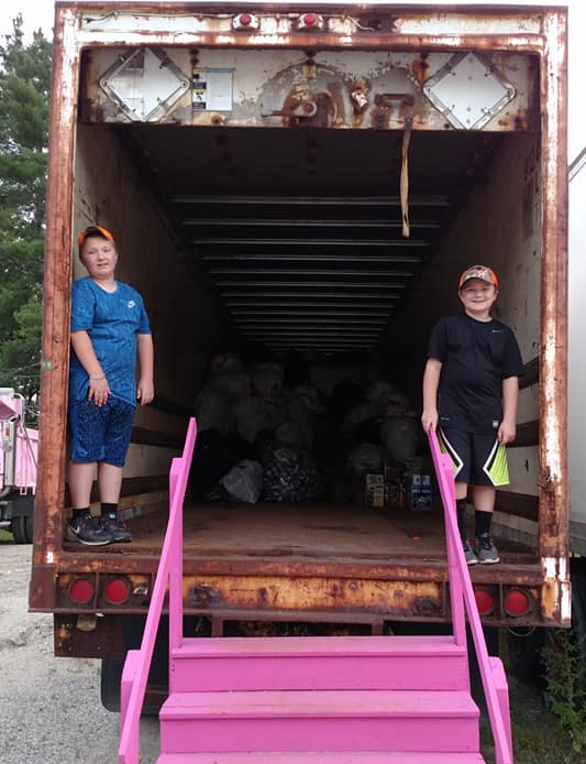 Two boys standing at the back of a rusted truck with pink stairs, filled with bags and boxes.