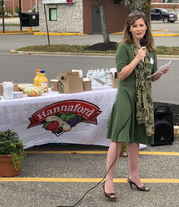 Woman speaking into a microphone at an outdoor event with a table displaying food and drinks in the background.