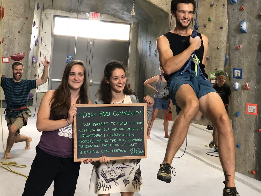 Group of people at indoor climbing gym holding sign about stewardship and conservation, with one person swinging on a rope.