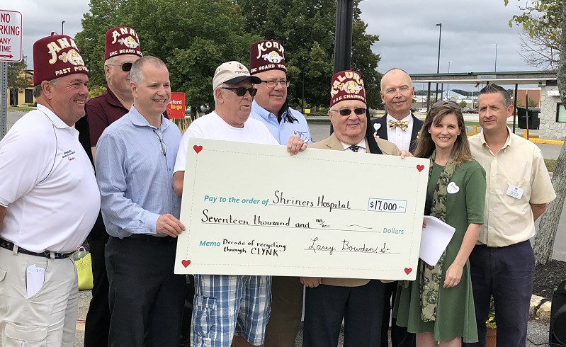 Group of people holding a large donation check for Shriners Hospital, with trees and a building in the background