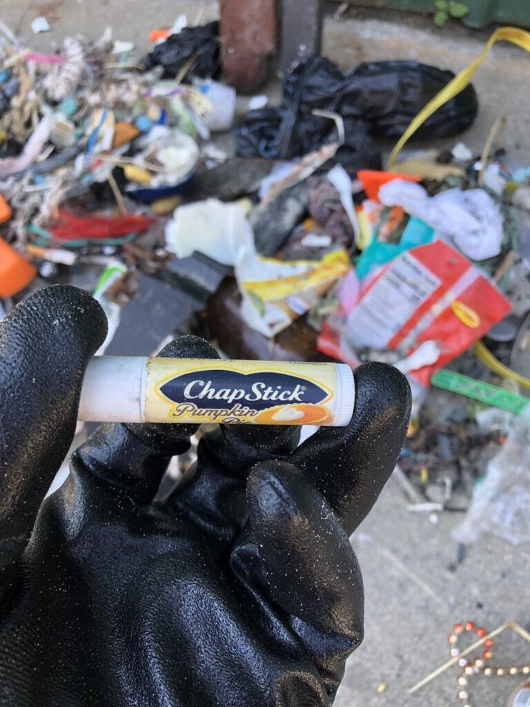 Person holding a ChapStick with pumpkin flavor among scattered trash and debris on the ground.