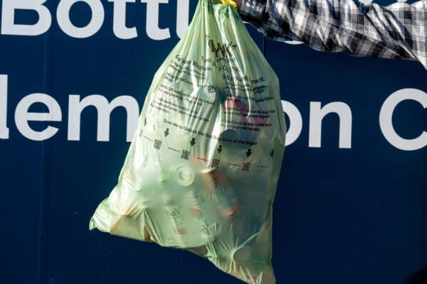 Person holding a green bag full of empty plastic bottles in front of a recycling information sign.