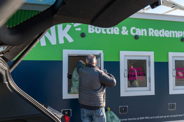 Person depositing bottles in a redemption machine at a recycling center parking lot, viewed through the trunk of a car.