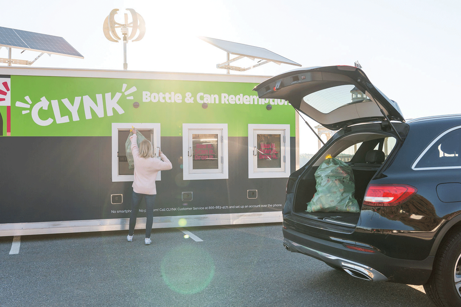 Person recycling bottles and cans at an outdoor redemption machine with an open car trunk filled with bags.