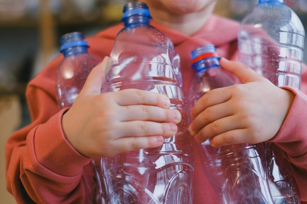 Child holding multiple empty plastic bottles, wearing a pink sweater, promoting recycling awareness and environmental responsibility.