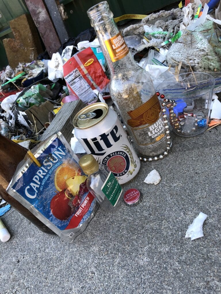 Litter scattered on ground: empty bottles, Capri Sun pouch, beer can, food packaging, and miscellaneous debris.