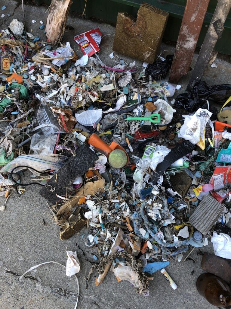 Pile of assorted garbage and debris on a concrete surface near a fence, including plastic, wood, and metal items.