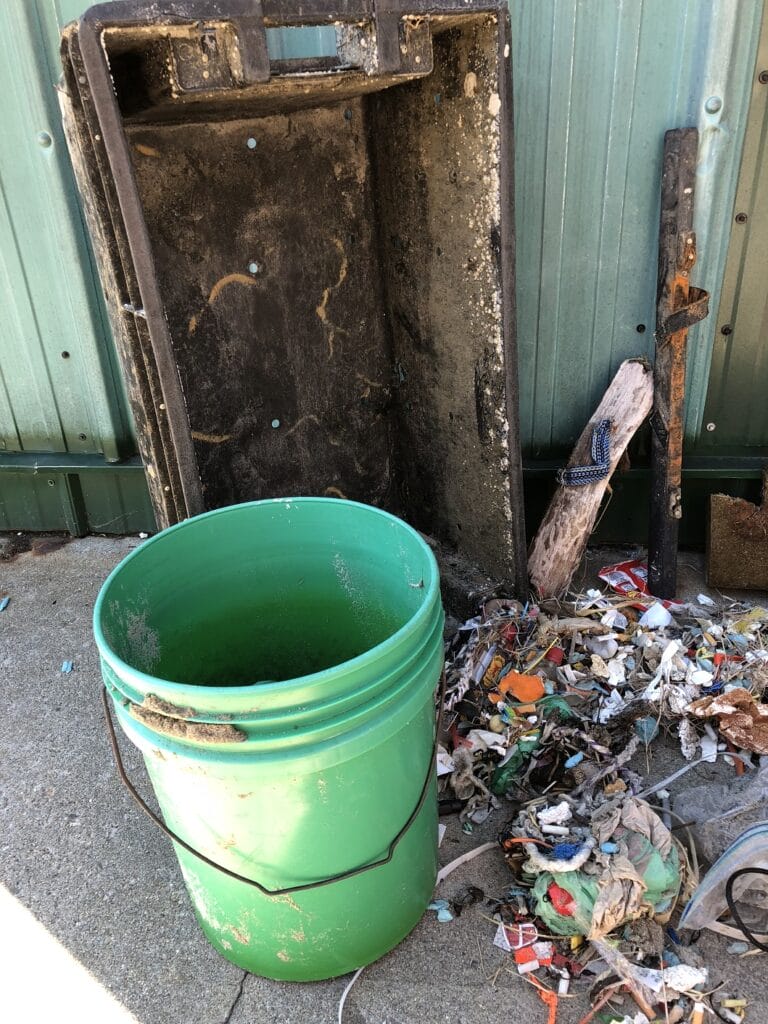 Green bucket and black container with scattered trash against a metal wall background outside on a concrete surface.