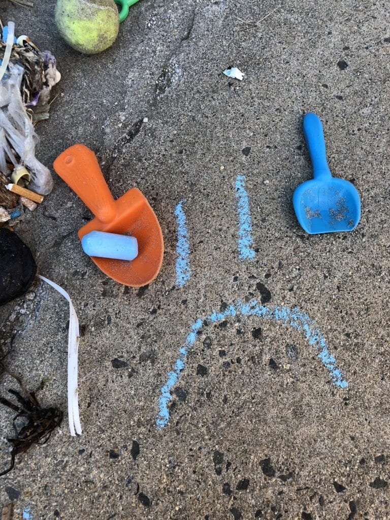 Two plastic shovels on concrete with a sad face drawn in blue chalk along with scattered debris and a worn-out tennis ball.