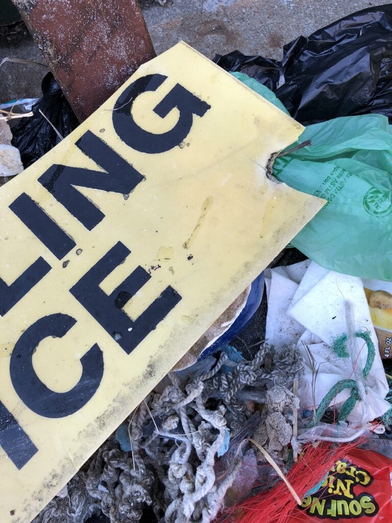 Damaged yellow sign with black text amid a pile of assorted trash and plastic bags.