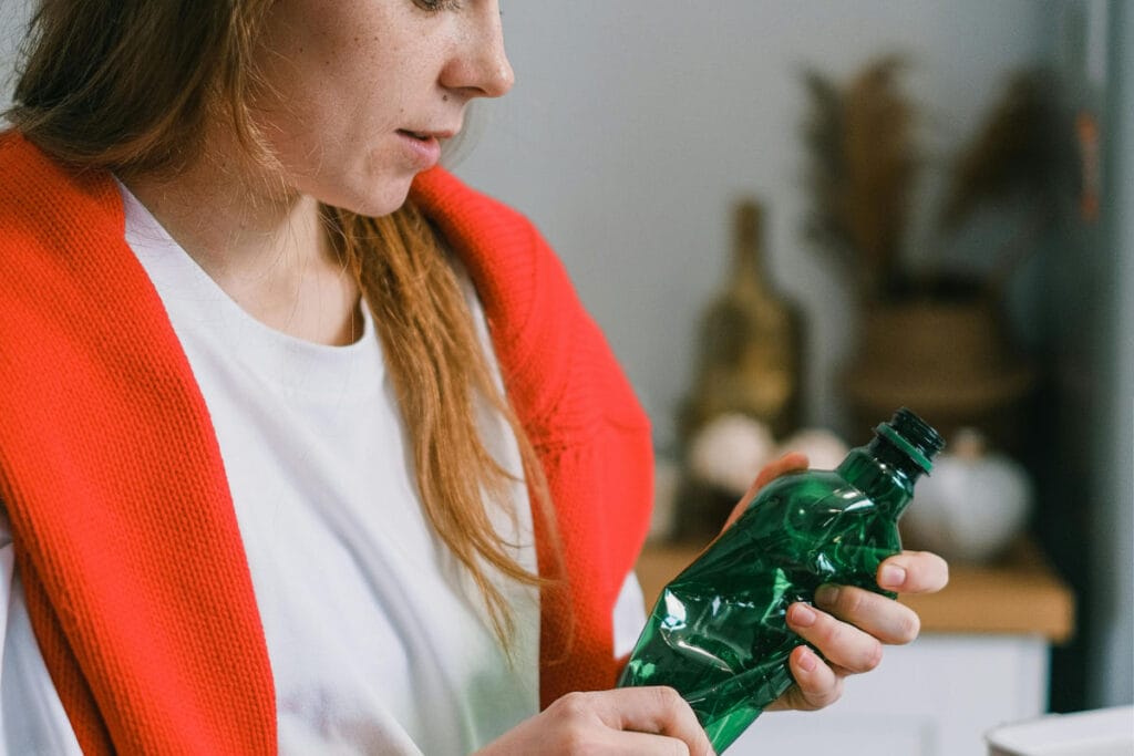 Person holding a crumpled green plastic bottle, wearing a red sweater and white shirt, indoors.