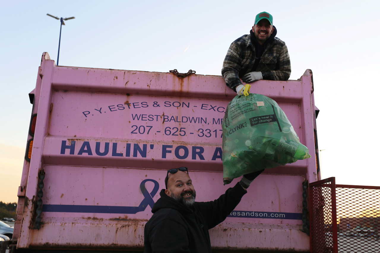 Two smiling men stand on and beside a pink garbage truck, one holding up a green bag of recyclables, sky in background.