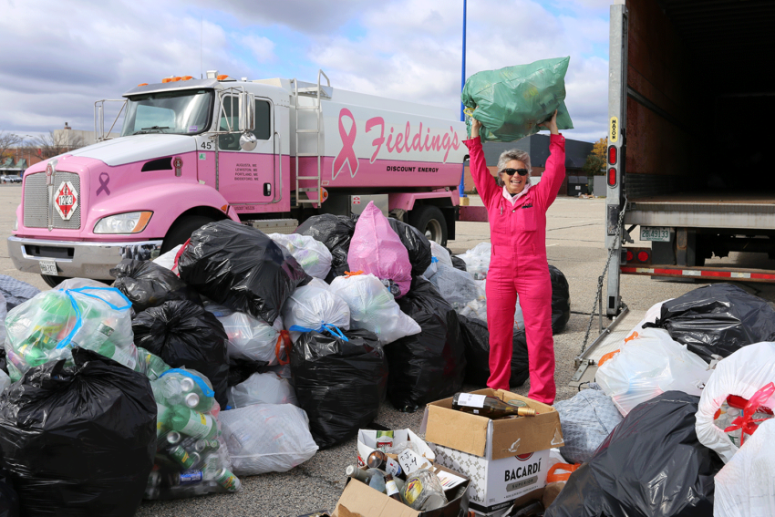 Person in pink standing next to a truck and piles of recycling bags, holds up a filled bag, smiling outdoors.