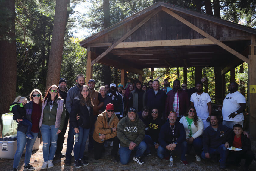 Group of people posing under a wooden shelter in a forest setting during a sunny day.