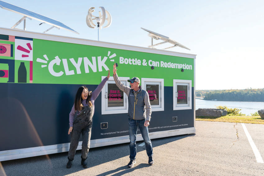 Two people high-fiving in front of a Clynk bottle and can redemption kiosk with solar panels.