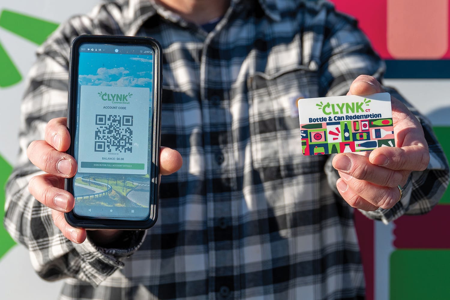 Person holding a smartphone displaying a QR code and a card for CLYNK bottle and can redemption program.