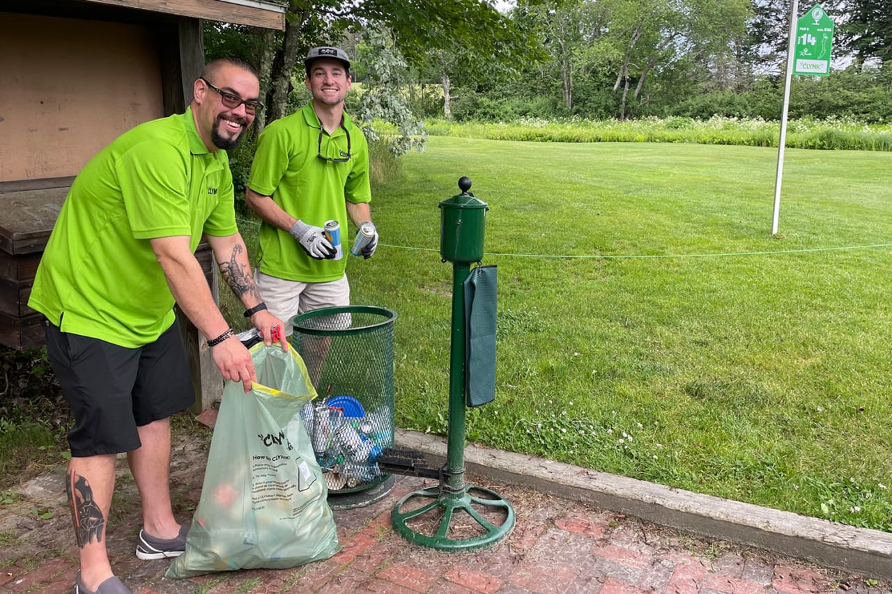 Two men in green shirts smiling while disposing of items into a trash bin on a sunny day in a park.