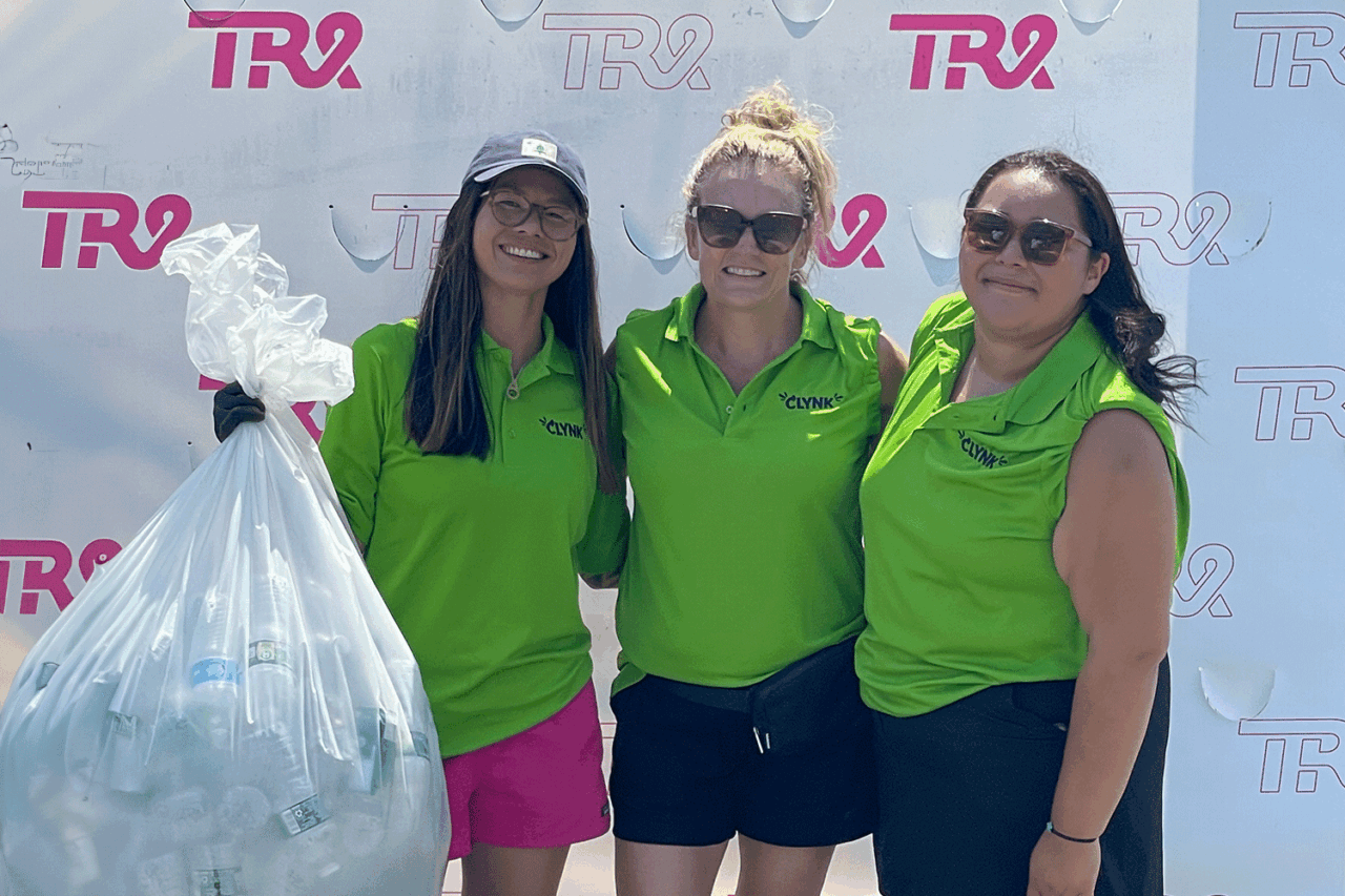 Three women in green shirts holding a bag of recyclables at a recycling event.