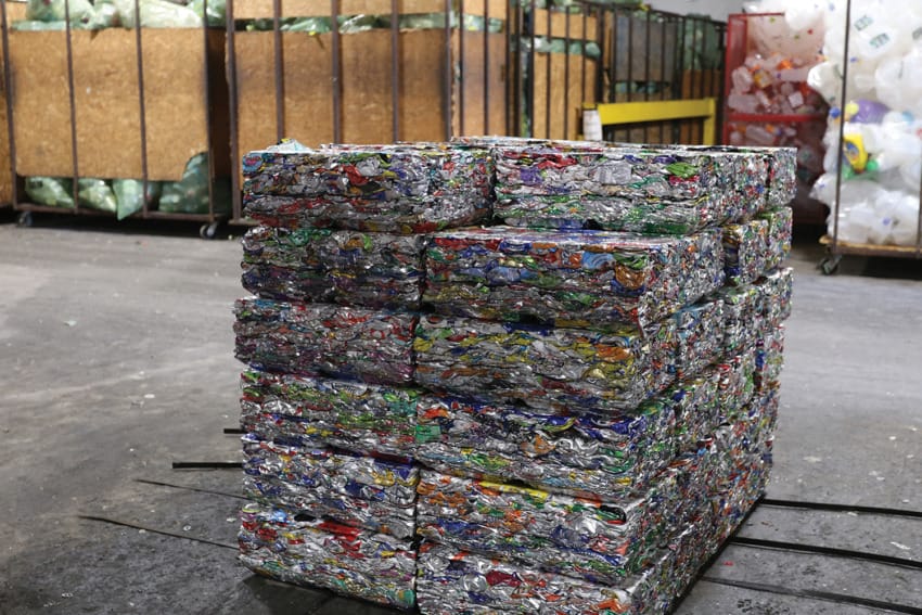 Stack of compacted aluminum cans in a recycling facility with sorted materials in the background.