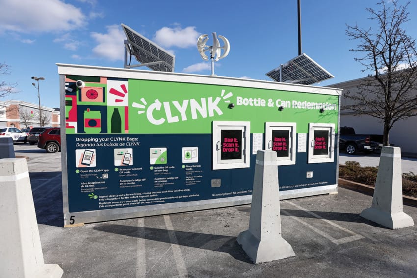 Bottle and can redemption kiosk with solar panels and instructional signs on a sunny day in a parking lot.