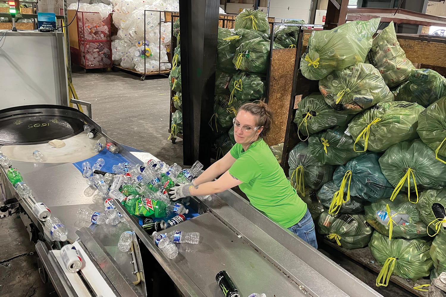 Woman sorting plastic bottles on recycling conveyor belt in facility surrounded by bags of recyclables.