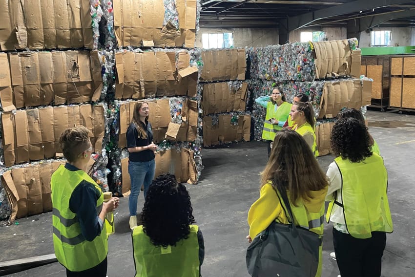 People in yellow safety vests touring a recycling facility with stacks of compressed materials in the background.