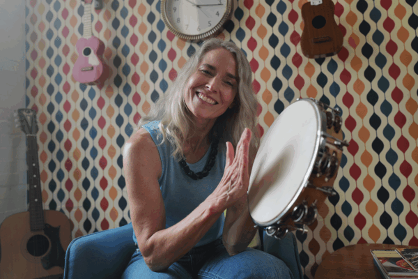 Smiling woman playing a tambourine seated in a room with colorful wallpaper and guitars on the wall.